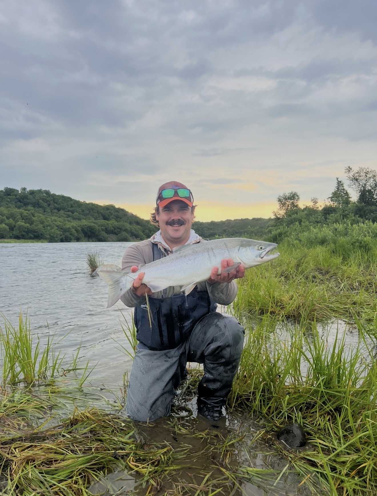 Captain Judd kneeling in the grass holding a bright silver salmon at sunset