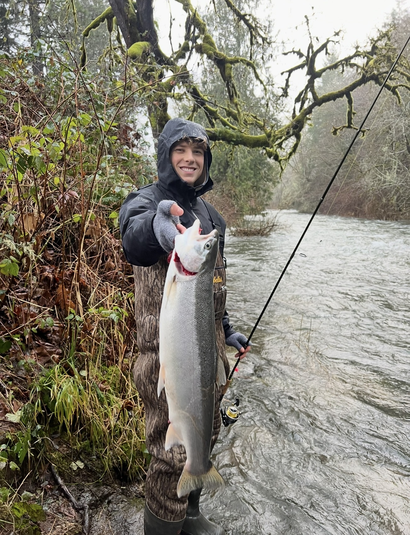 Young angler smiling with a chrome winter steelhead on a mossy Oregon river
